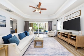 A living room with a ceiling fan, a sofa with blue cushions, a coffee table, a TV on a wooden stand, and a view of the outdoors through large windows. - Last Minute Disney Getaway! Luxury Condo with Private Balcony. Book Now! #87723 (Orlando)
