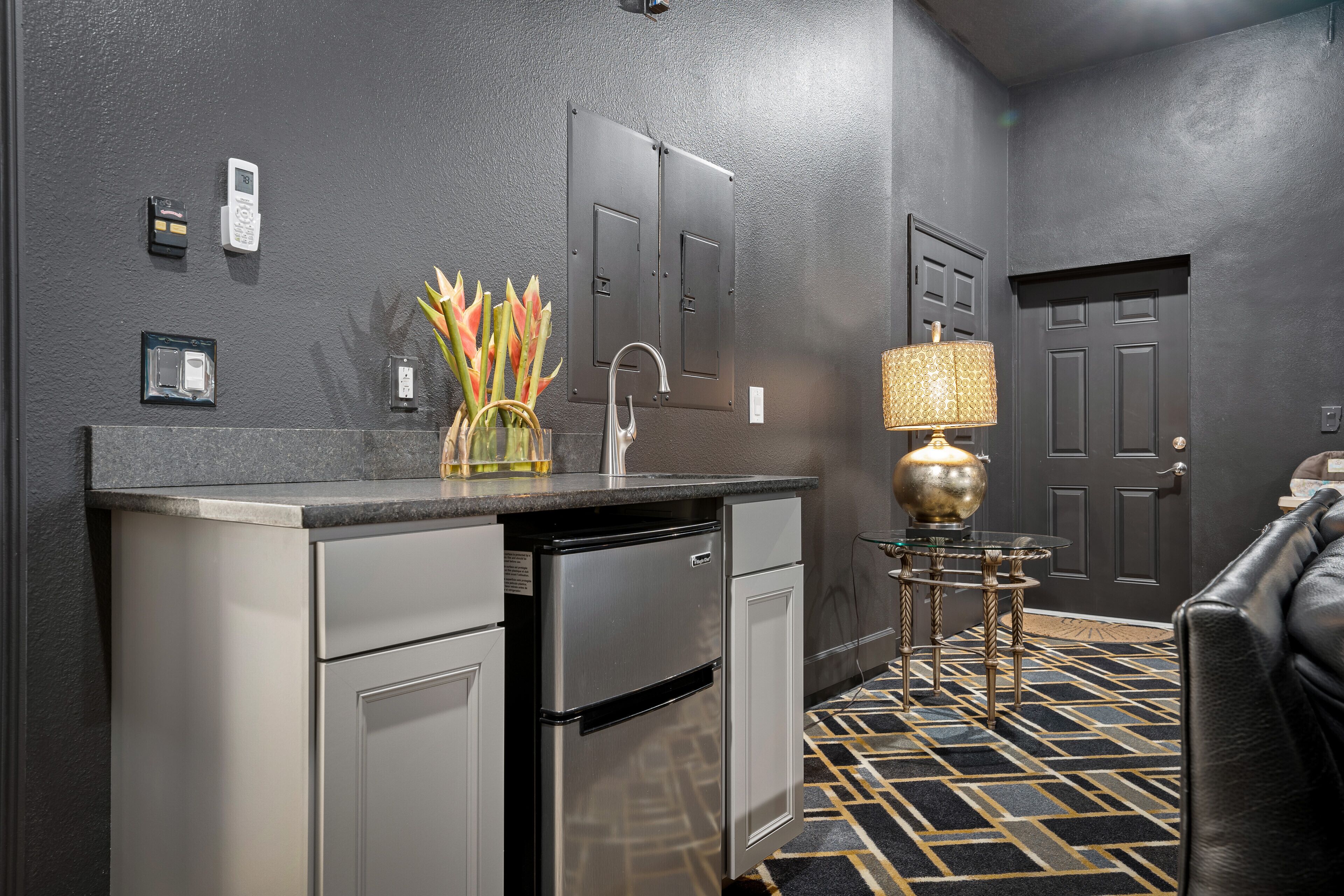 A compact kitchen with a built-in refrigerator, a sink, and a small countertop. There is a decorative vase with flowers on the counter, and a lamp on a small table. The floor has a geometric patterned tile design.