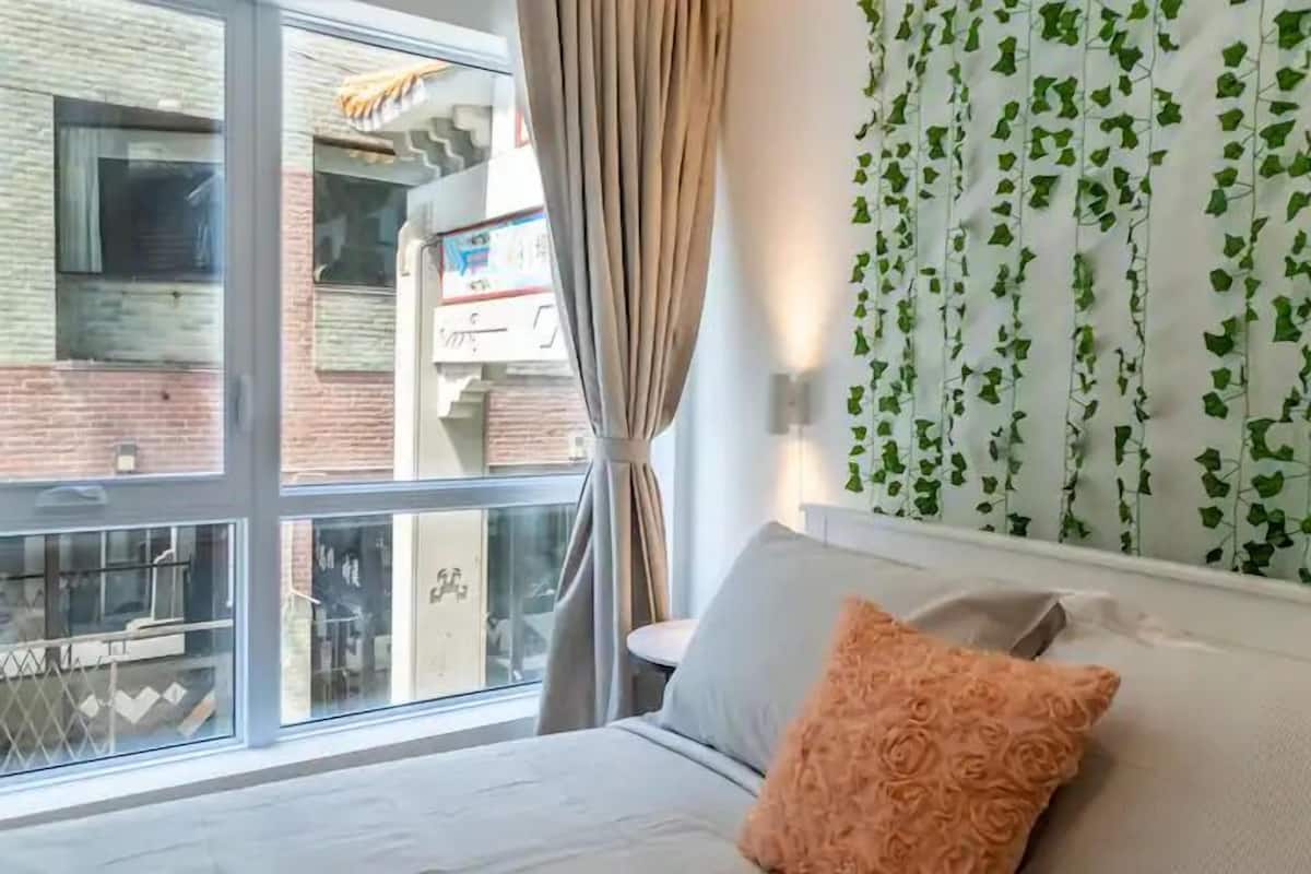Une chambre avec une grande fenêtre, un lit avec un oreiller de couleur pêche et un mur recouvert de lierre vert.