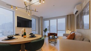 Uma sala de estar moderna com sofá, mesa de jantar com cadeiras, televisão na parede e grandes janelas com vista para a cidade.