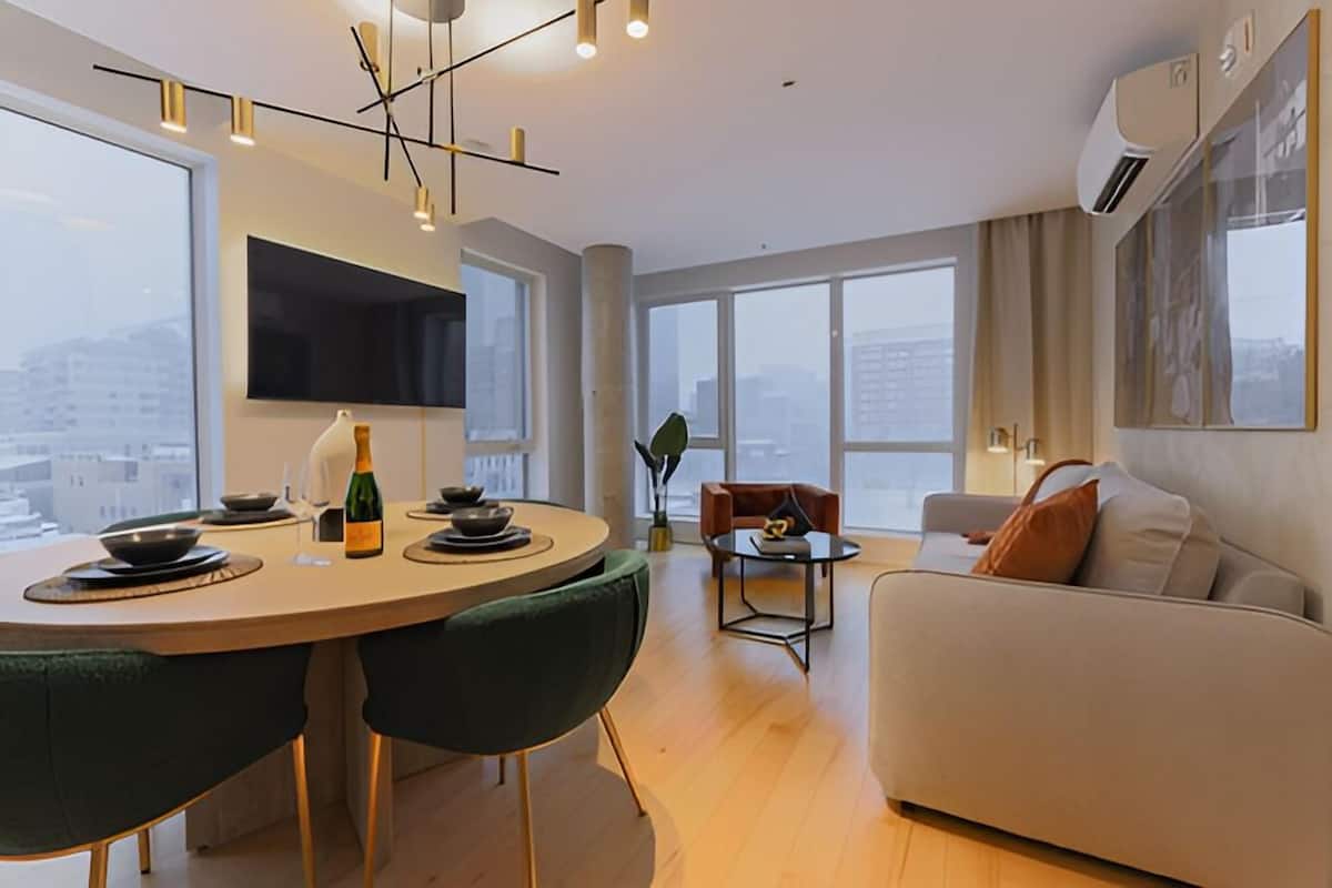 Un salon moderne comprenant un canapé, une table à manger avec des chaises, un téléviseur fixé au mur et de grandes fenêtres offrant une vue sur la ville.