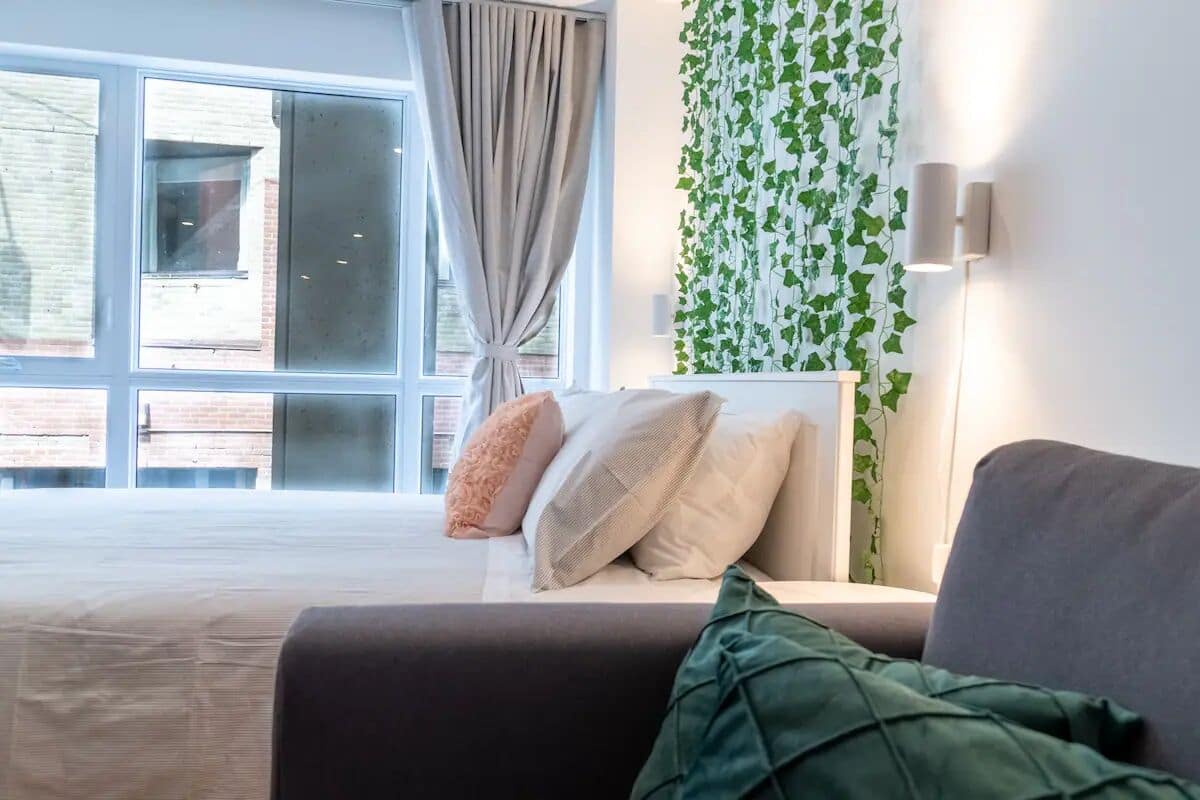 Une chambre moderne avec une grande fenêtre, un lit avec une literie blanche, un canapé gris et un plaid vert.