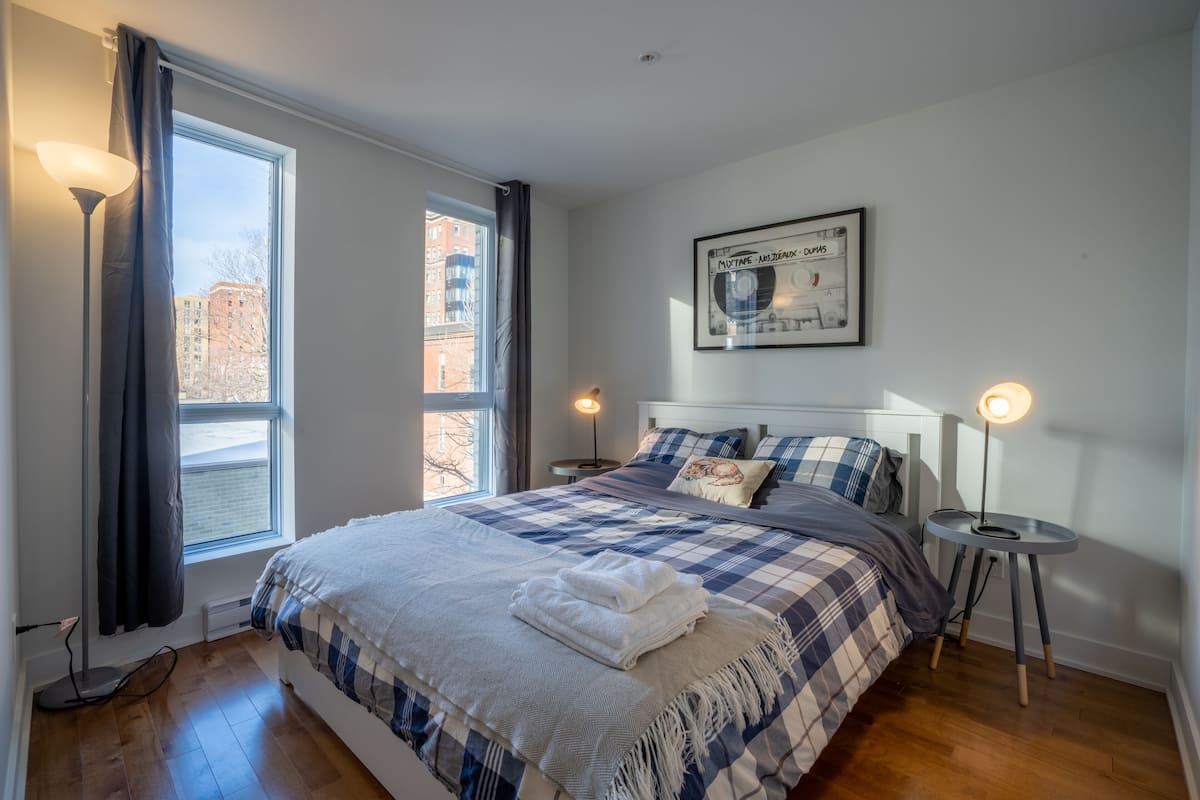 Une chambre avec un grand lit, deux tables de chevet, une fenêtre avec des rideaux, une œuvre d’art accrochée au mur et une lampe sur pied.