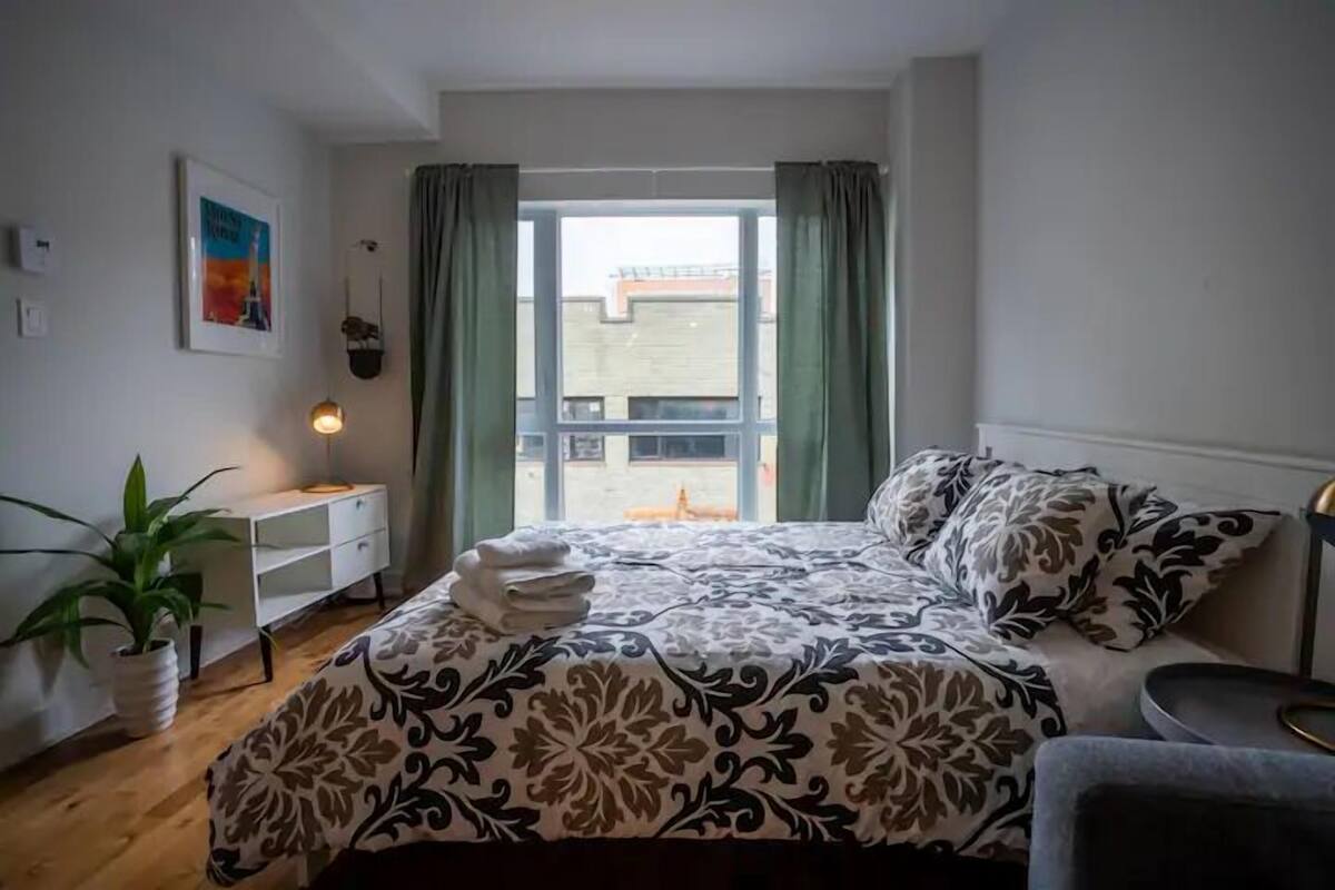 Une chambre à coucher avec un lit, une table de chevet, une lampe, une plante en pot et une fenêtre avec des rideaux.