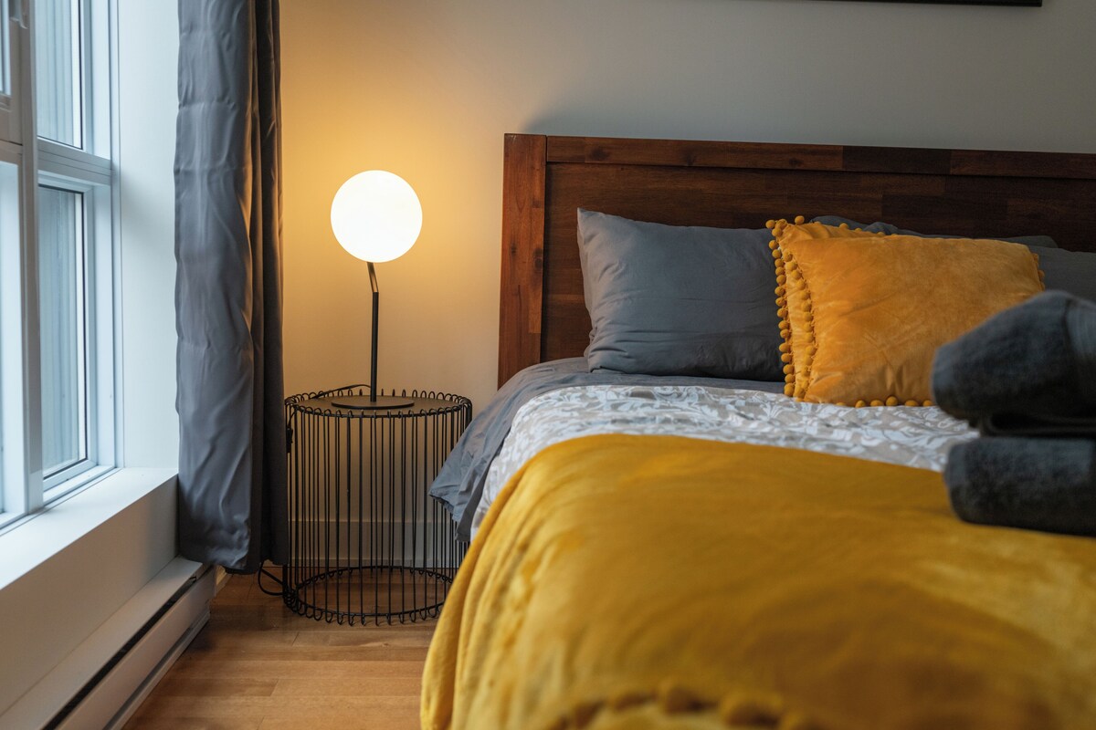 A bedroom with a wooden headboard, a bedside lamp, a window with curtains, and a bed with a yellow blanket and pillows.