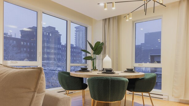 Uma área de refeições moderna com uma mesa redonda, cadeiras estofadas em verde e vista para prédios da cidade através de grandes janelas.