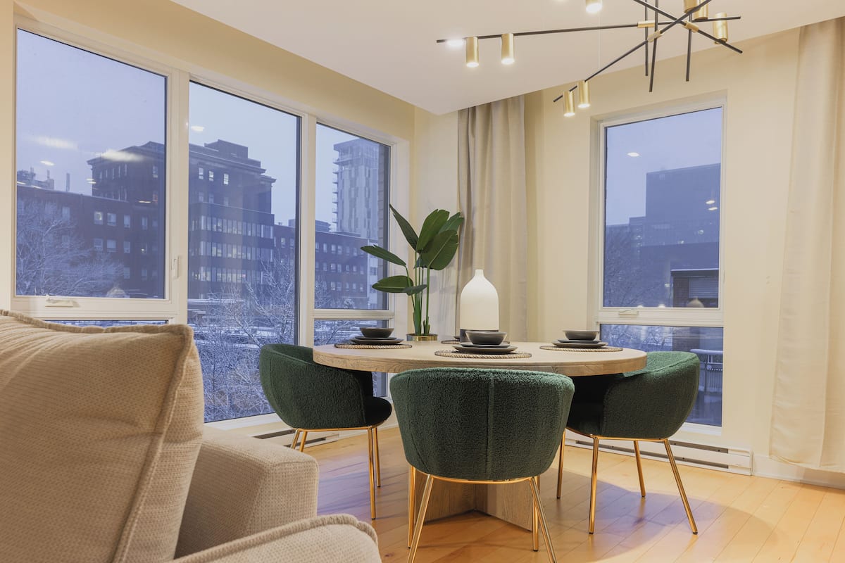 Un espace repas moderne avec une table ronde, des chaises recouvertes d’un tissu vert et une vue sur les bâtiments de la ville, grâce à de grandes fenêtres.