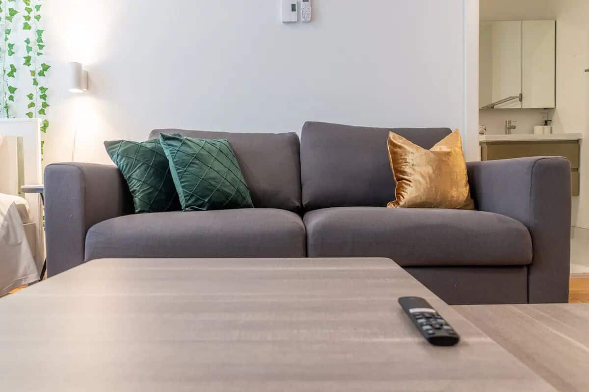 Un canapé gris avec deux coussins, une télécommande posée sur une table en bois, et un mur blanc avec un interrupteur.