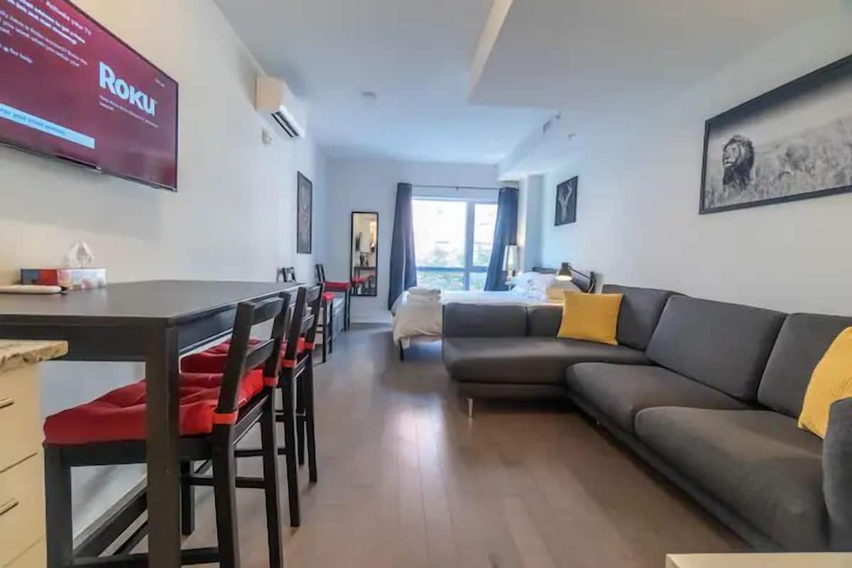 Un salon moderne avec un canapé gris, une table à manger avec des chaises rouges, un téléviseur fixé au mur et une grande fenêtre avec des rideaux.