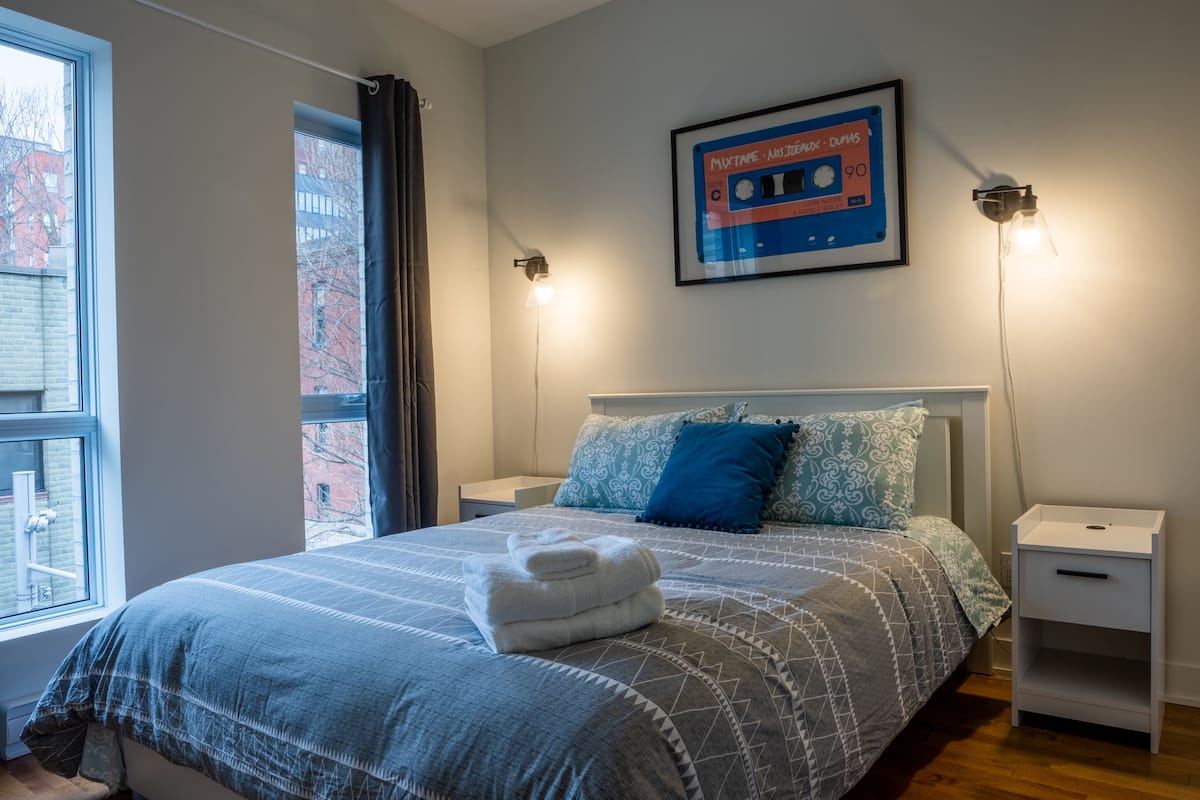 Un lit bien fait, recouvert d’une couvre-lit à motifs, deux tables de chevet avec des lampes, un tableau encadré au mur et une fenêtre avec des rideaux.