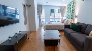 Quarto de hotel moderno com cama, sofá, televisão, mesa pequena e janela com cortinas.