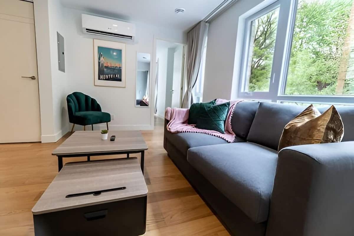 Un salon moderne avec un canapé gris, un fauteuil vert, une table basse et une grande fenêtre donnant sur des arbres.