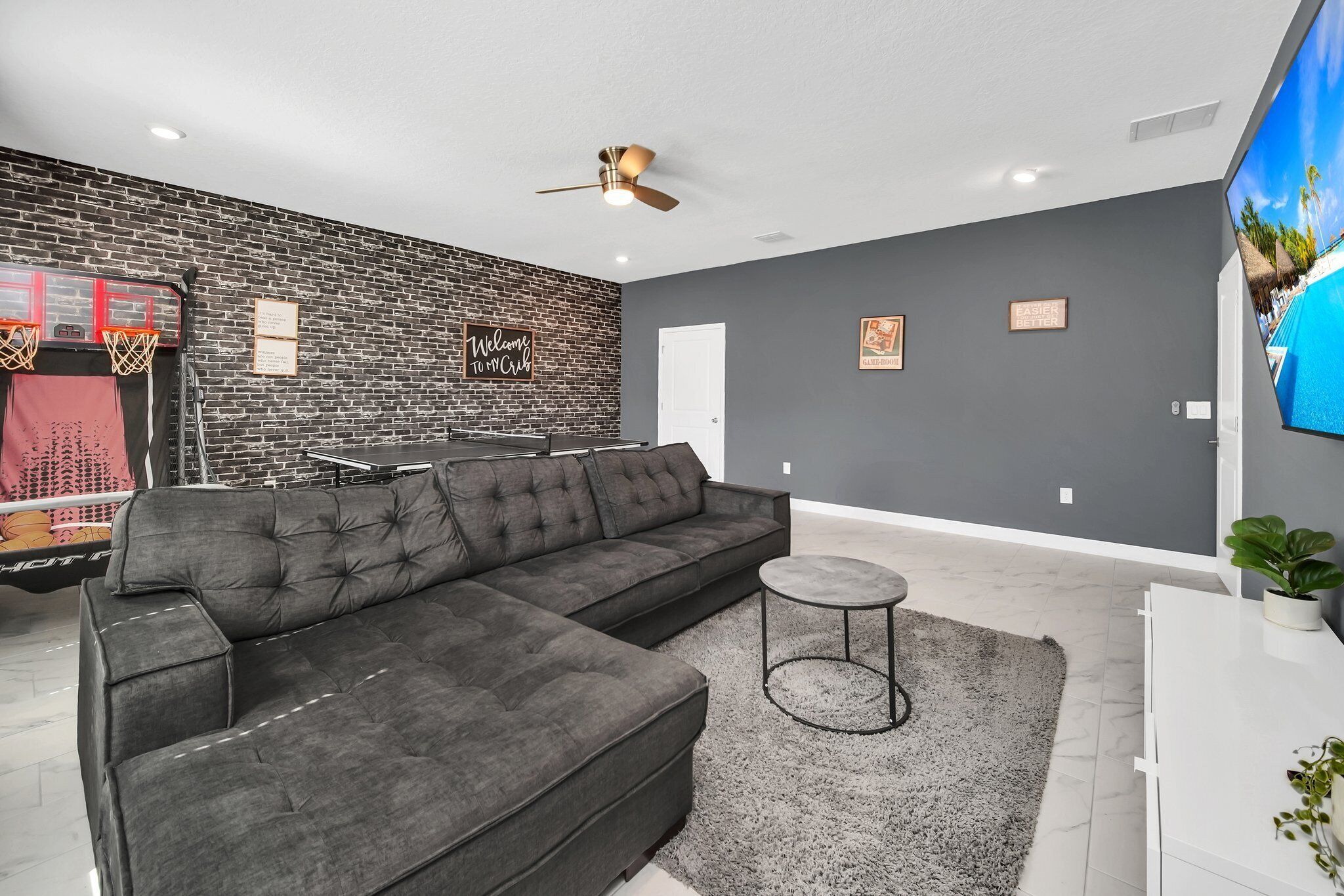 A modern living room with a sectional sofa, a small round table, a brick accent wall, a ceiling fan, and a TV displaying a pool scene.