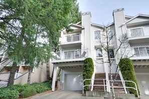 Un immeuble résidentiel à plusieurs étages, doté d’un garage central, d’un escalier menant aux étages supérieurs et d’un jardin bien entretenu avec des arbustes taillés et un arbre.