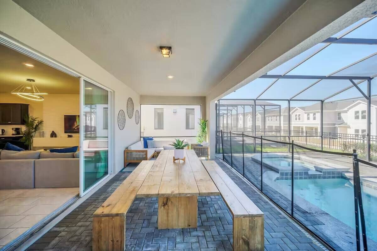 A modern living room with a wooden table and benches, a sofa, and a glass door leading to a balcony with a pool.