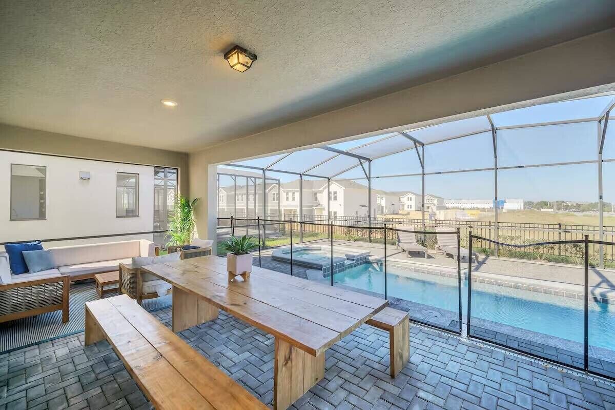 A poolside area with a wooden table and benches, a glass-enclosed pool, and a view of surrounding buildings.