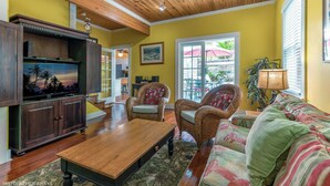 A living room with a wooden ceiling, a television, wicker furniture, and a view of the outside. - Truman House (Key West)