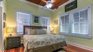 A bedroom with a wooden bed, two bedside tables with lamps, a ceiling fan, and large windows with blinds. - Truman House (Key West)