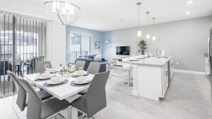 Cozinha moderna com ilha central, área de refeições com mesa branca posta e sala de estar com sofá azul.