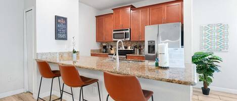 Una cocina moderna con encimera de granito, electrodomésticos de acero inoxidable y armarios de madera. Hay tres taburetes de bar y una planta en maceta.