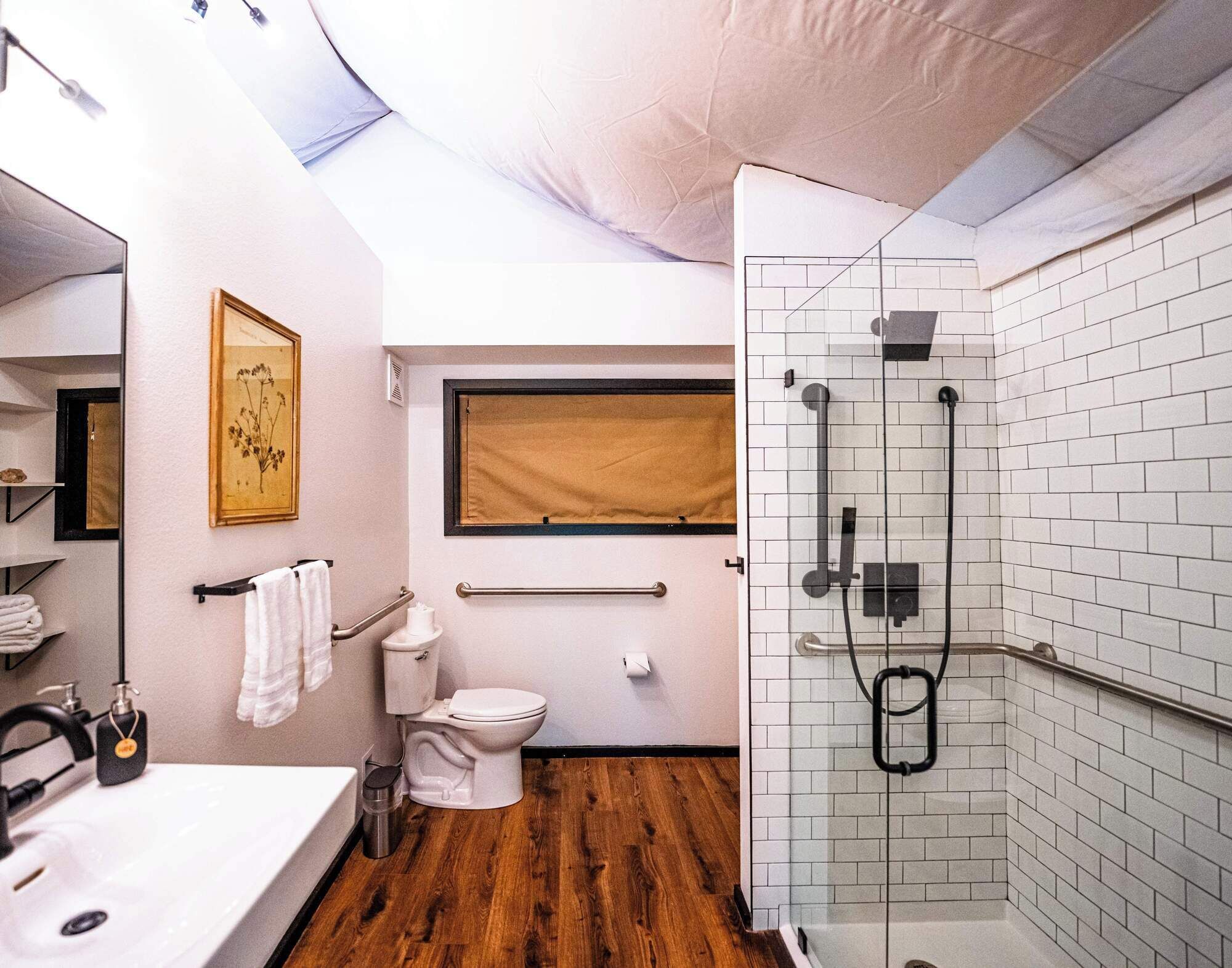 A modern bathroom with a wooden floor, white fixtures, a glass shower enclosure, and a window with a beige blind.
