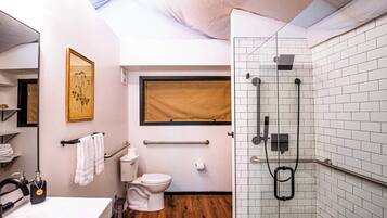 A modern bathroom with a wooden floor, white fixtures, a glass shower enclosure, and a window with a beige blind.
