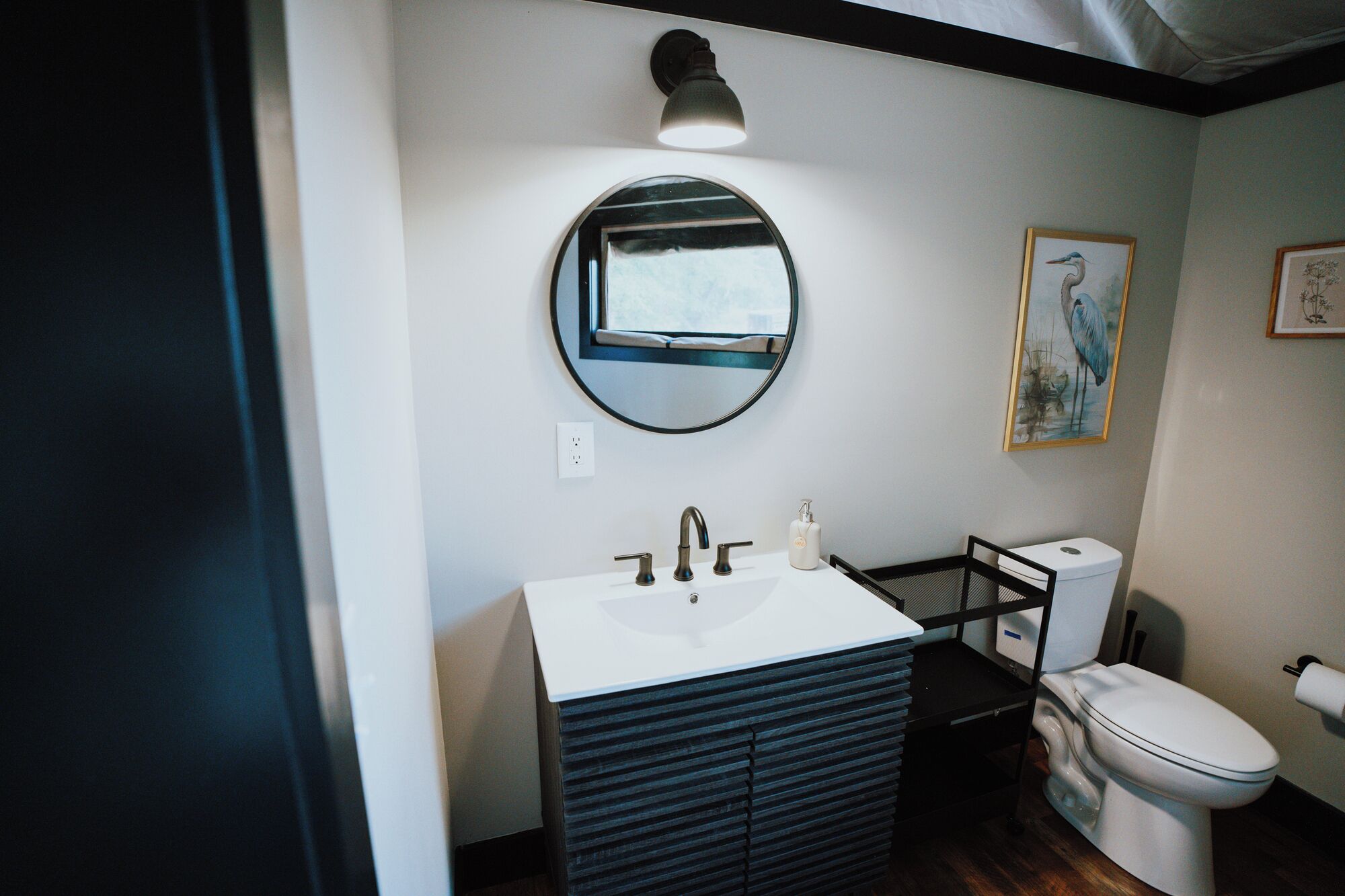 A bathroom with a round mirror, a white sink, a black cabinet, a toilet, and a framed artwork on the wall.