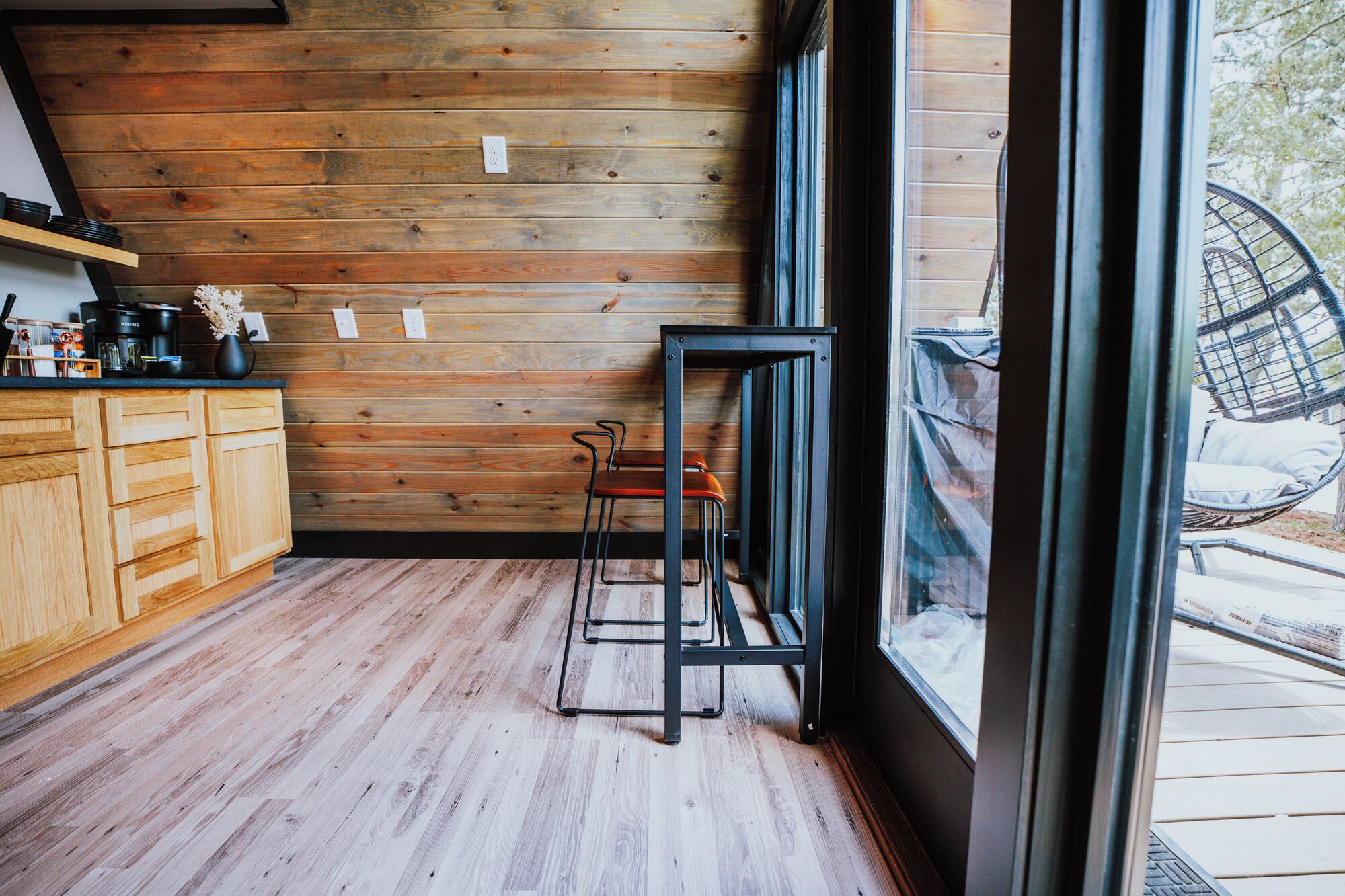 A kitchen with wooden walls, a black metal stool, and a window with a view of the outdoors.