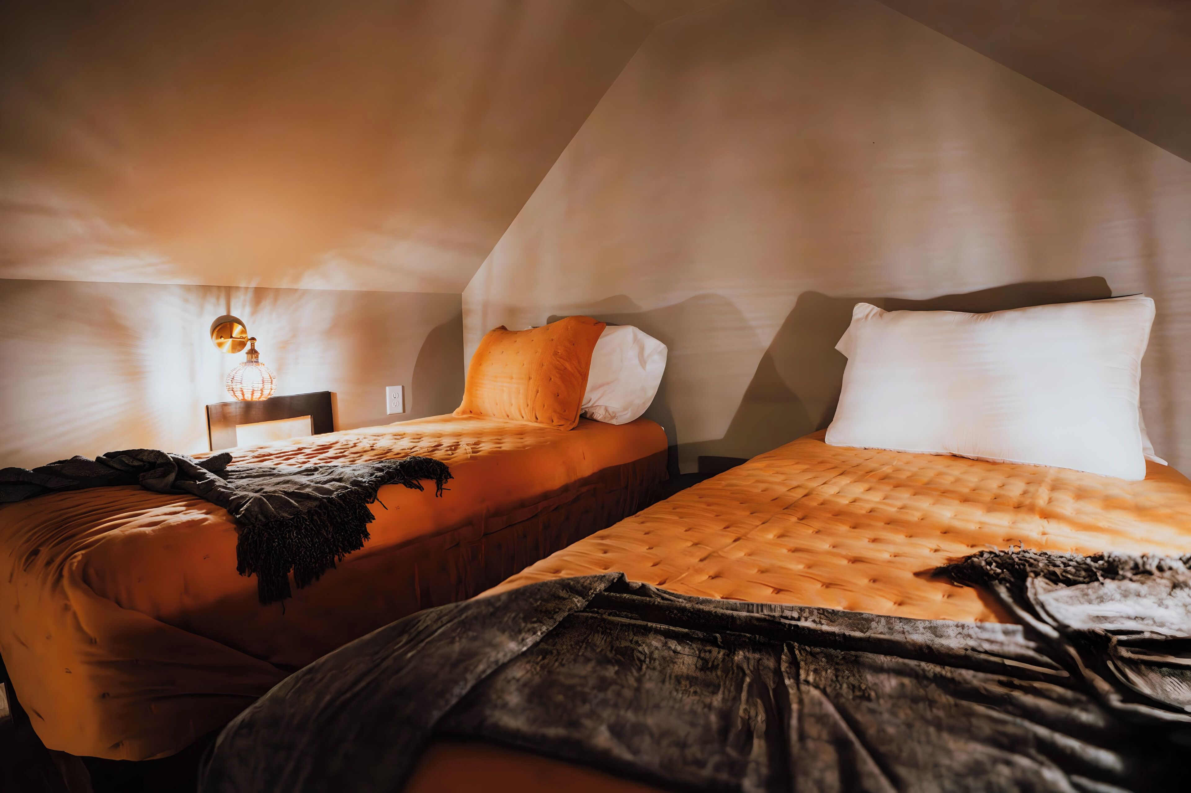 Two beds with orange bedding, a wooden headboard, and a hanging light fixture in a room with sloped ceilings.