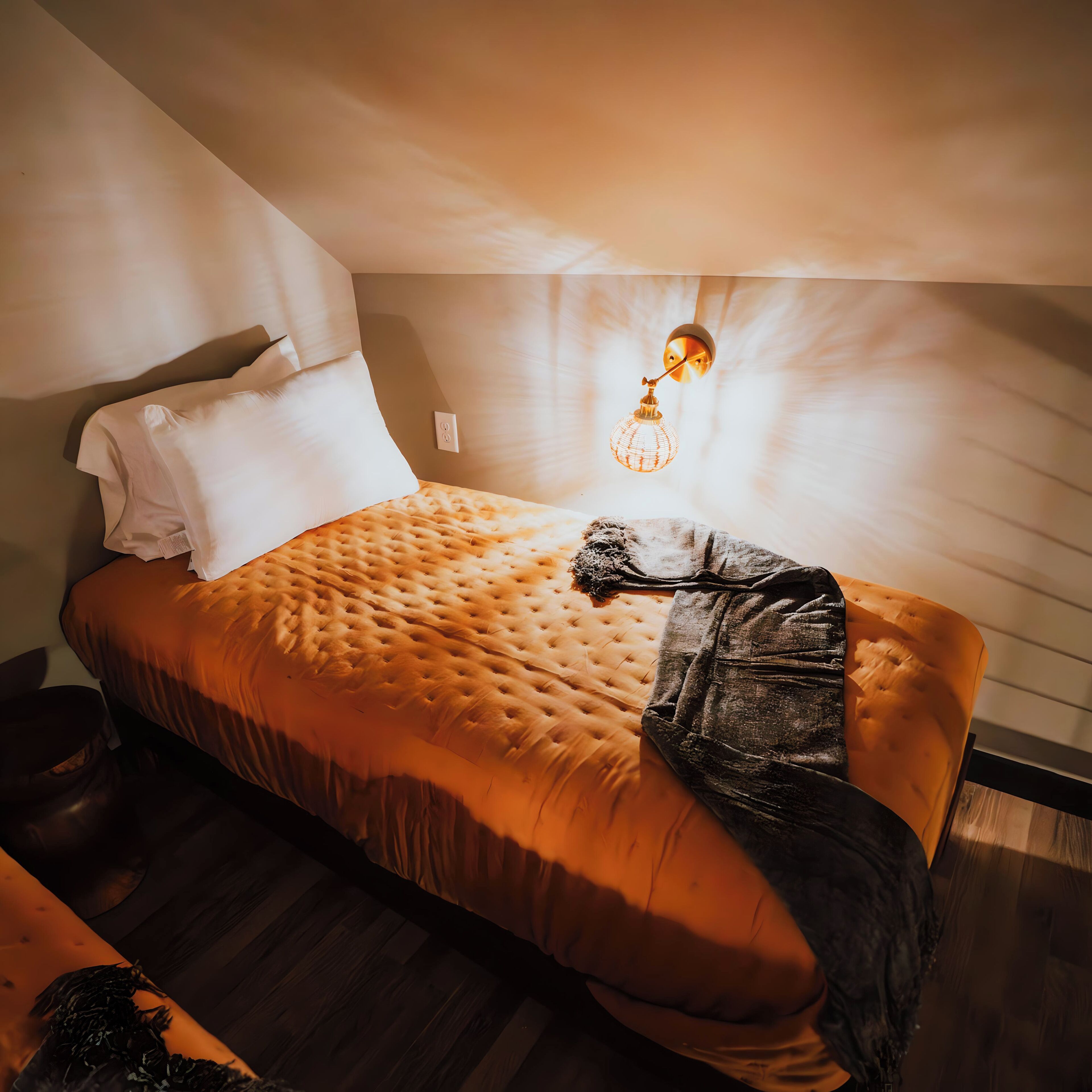 A neatly made bed with a tufted headboard, a single pillow, and a blanket draped over the side. A unique wall-mounted light fixture is visible above the bed.