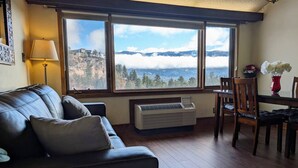 A living room with a dark leather sofa, a table with chairs, a window with a view of mountains and clouds, and a standing heater.