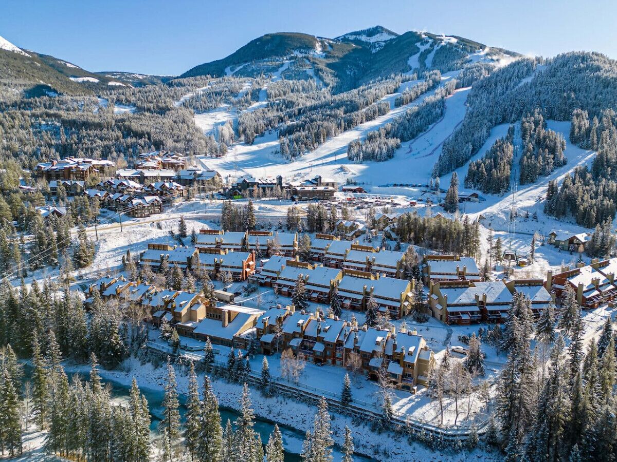 Une station de ski enneigée, avec plusieurs bâtiments, des pistes de ski et, en arrière-plan, des montagnes recouvertes de forêts.