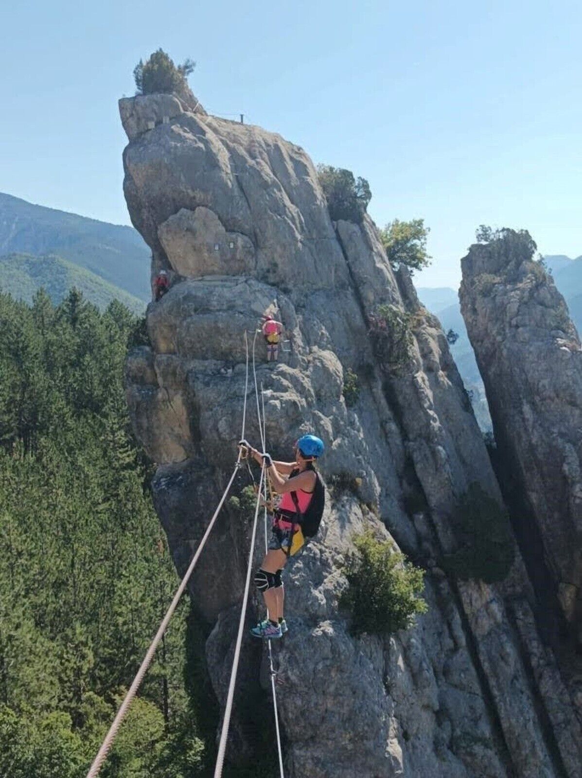 A person rappelling down a rocky cliff with a harness and helmet, surrounded by forested mountains.