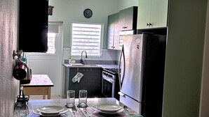 Uma cozinha compacta com pia, fogão e geladeira. Há uma janela com persianas, um relógio na parede e uma mesa posta com pratos e talheres.