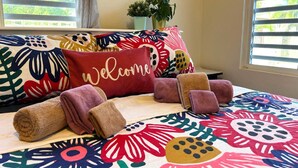 Cama com roupa de cama floral colorida, um travesseiro com a palavra 'Welcome' e toalhas dobradas.