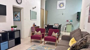 Uma sala de estar com sofá, duas poltronas, mesa de centro, televisão, mesa de jantar com cadeiras, um pequeno armário e um quadro na parede.