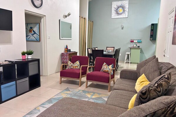A living room with a sofa, two armchairs, a coffee table, a TV, a dining table with chairs, a small cabinet, and a painting on the wall.
