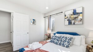 Un dormitorio ordenado con una cama, una puerta blanca, un cuadro y una ventana con cortinas.