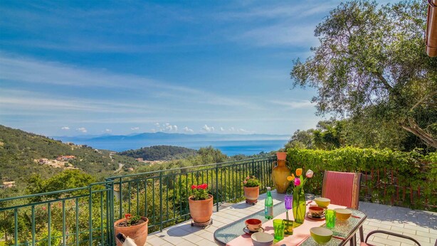 Une terrasse avec une table dressée pour le repas, des plantes en pot et une vue panoramique sur la mer et les collines.