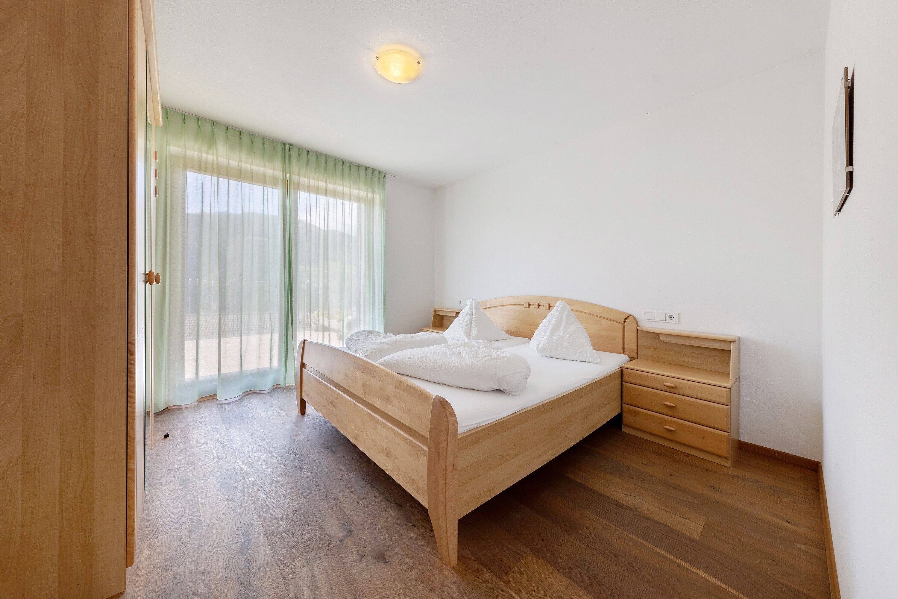 A bedroom with a wooden bed, a nightstand, a dresser, and a window with sheer curtains.