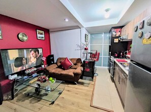 Un espace de vie compact comprenant un canapé, un téléviseur, une table basse en verre et une kitchenette.
