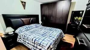 Ein Schlafzimmer mit Bett, Nachttisch, Kommode, Spiegel und einem an der Wand hängenden Kruzifix.