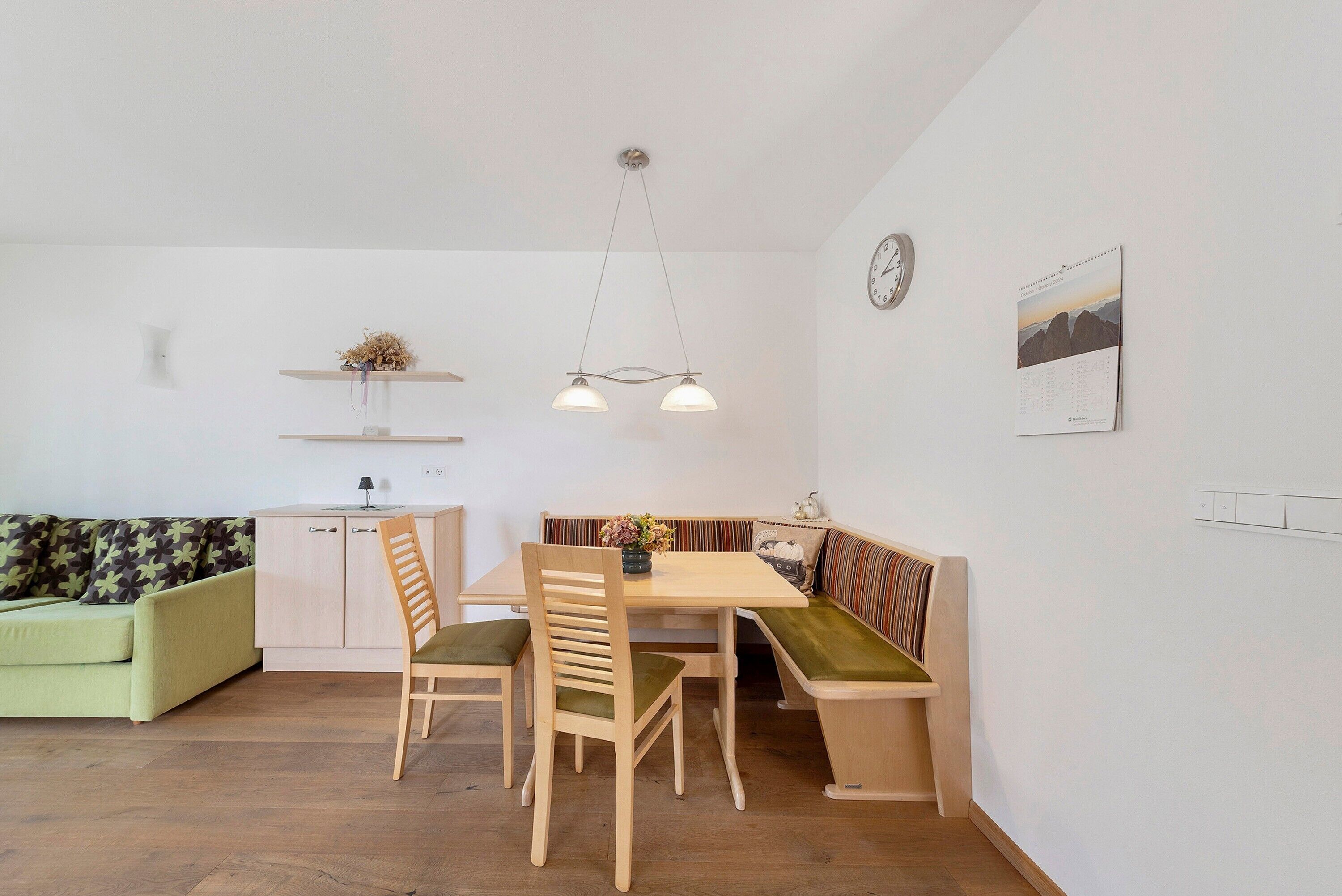 A dining area with a wooden table, chairs, and a bench. There is a light fixture above the table and a clock on the wall.
