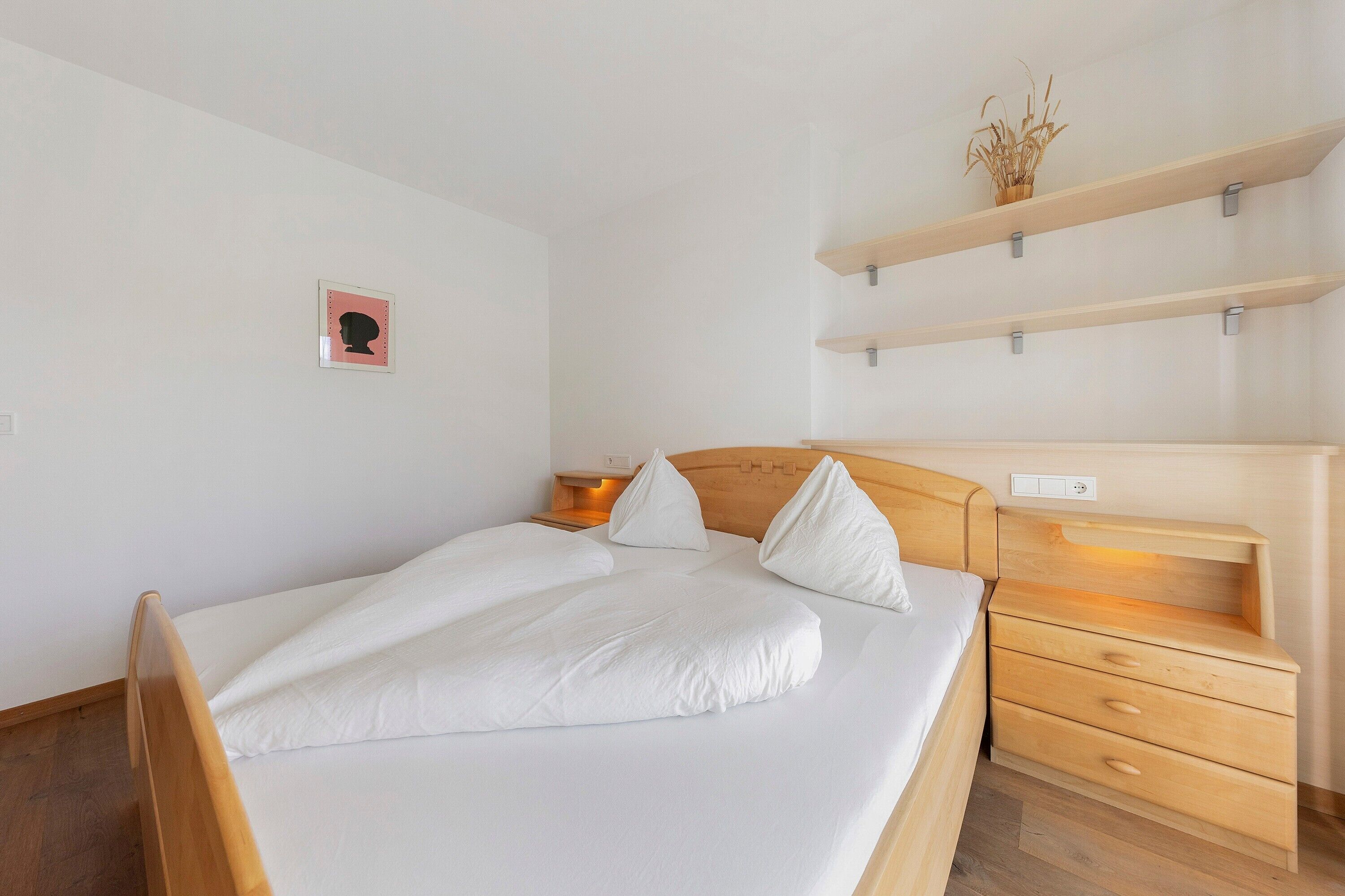 Una cama bien hecha con sábanas blancas, un cabecero de madera y una mesita de noche con cajones. Encima de la cama, hay un pequeño estante con una planta en la pared.