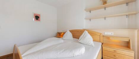 Una cama bien hecha con sábanas blancas, un cabecero de madera y una mesita de noche con cajones. Encima de la cama, hay un pequeño estante con una planta en la pared.
