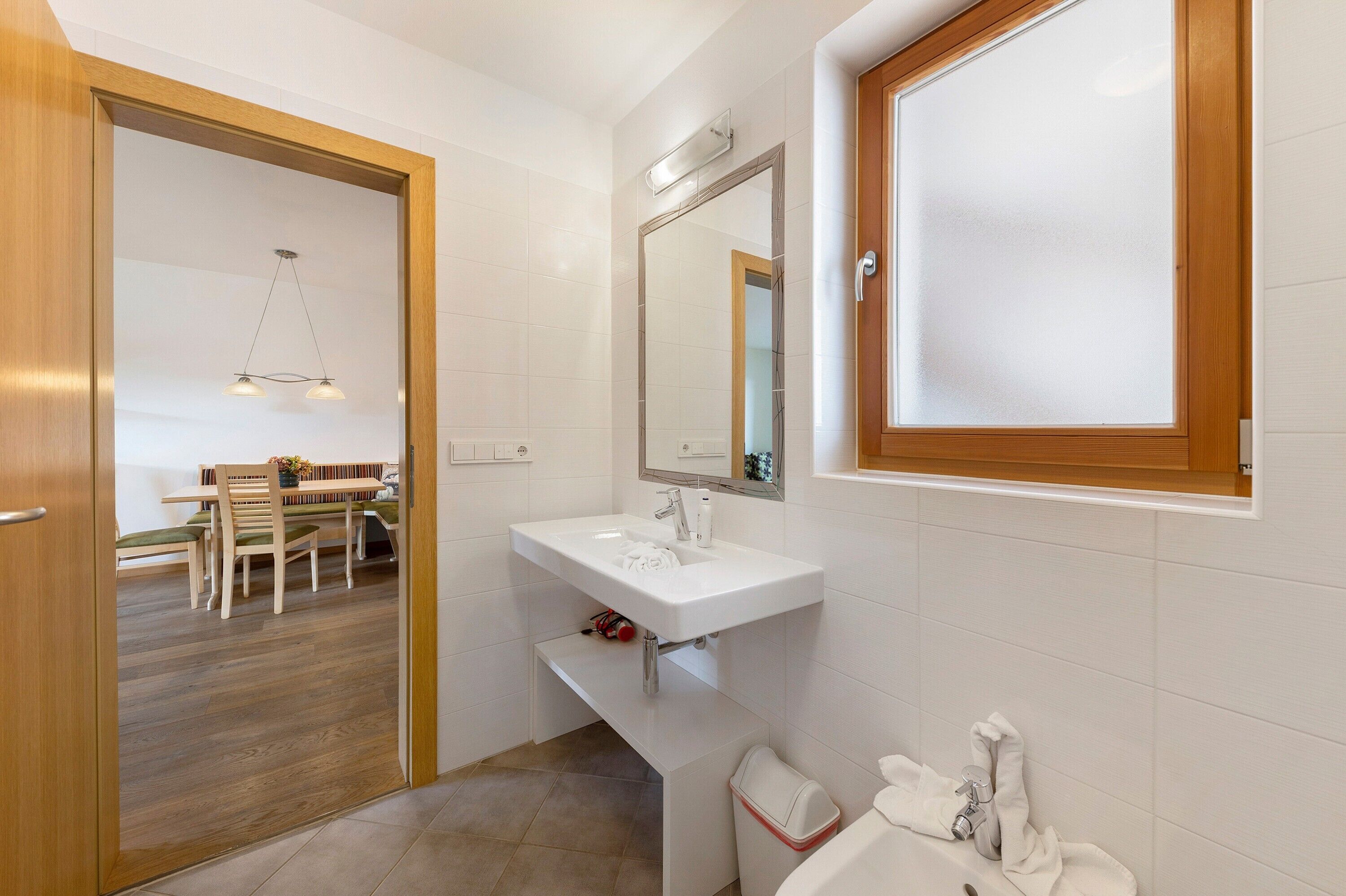 A bathroom with a white sink, mirror, and toilet. There is a wooden door leading to a dining area with a table and chairs.