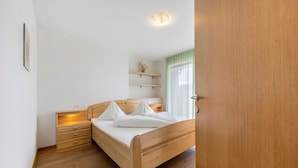 A bedroom with a wooden bed, bedside tables, a nightstand, a wall-mounted shelf with a plant, and a window with curtains.