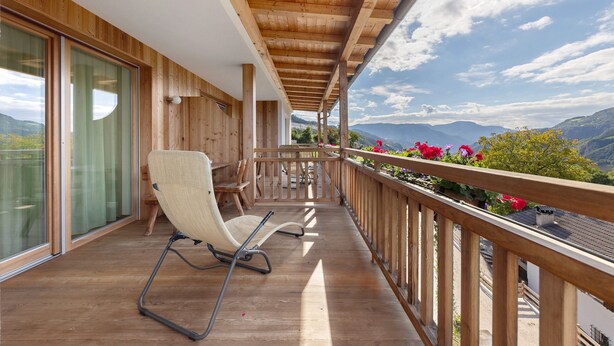 Un balcón de madera con una silla, una mesa y vistas a montañas.