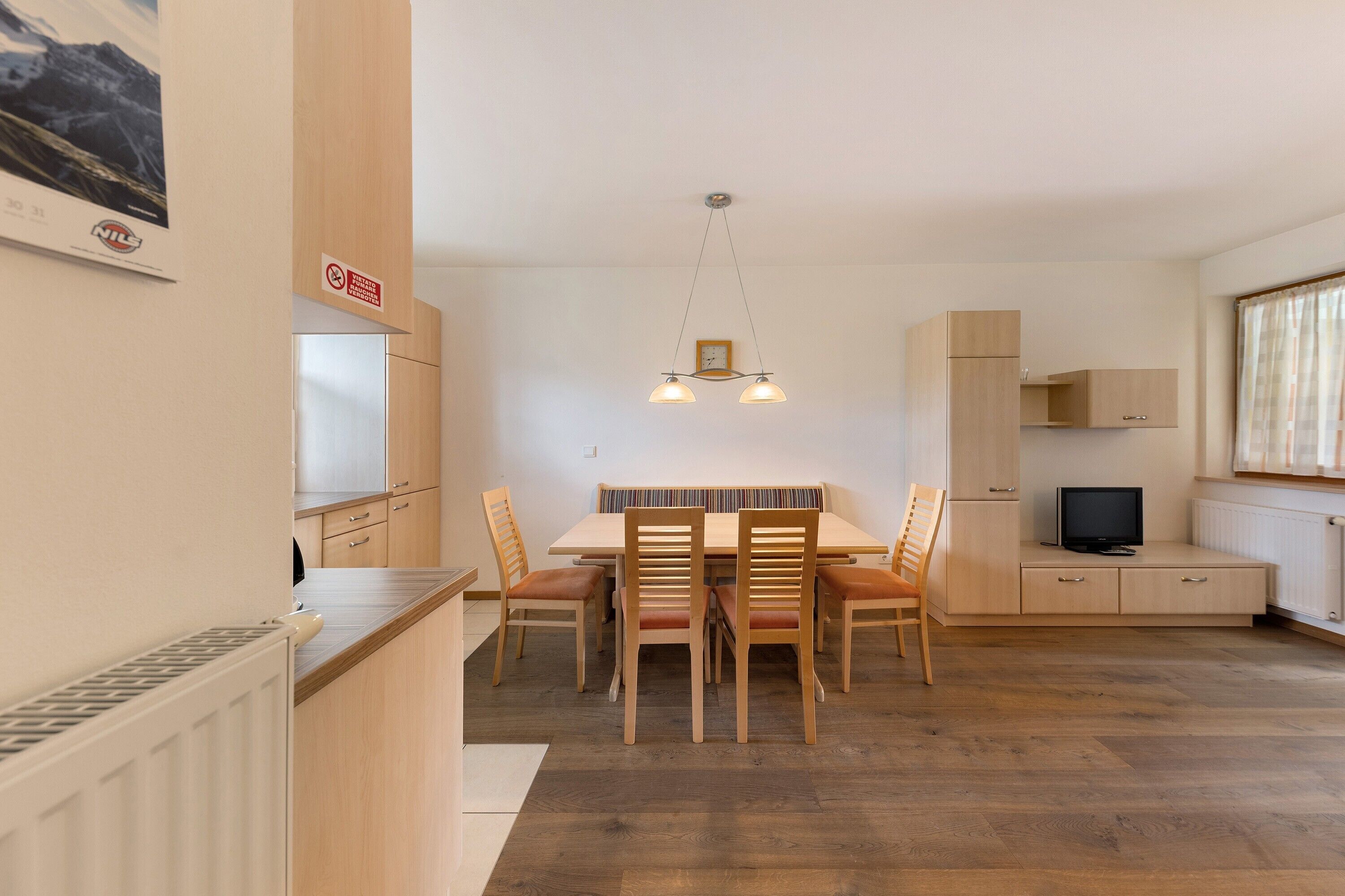 Une cuisine moderne avec des meubles en bois, une table à manger avec des chaises, un luminaire suspendu et un téléviseur posé sur un meuble.