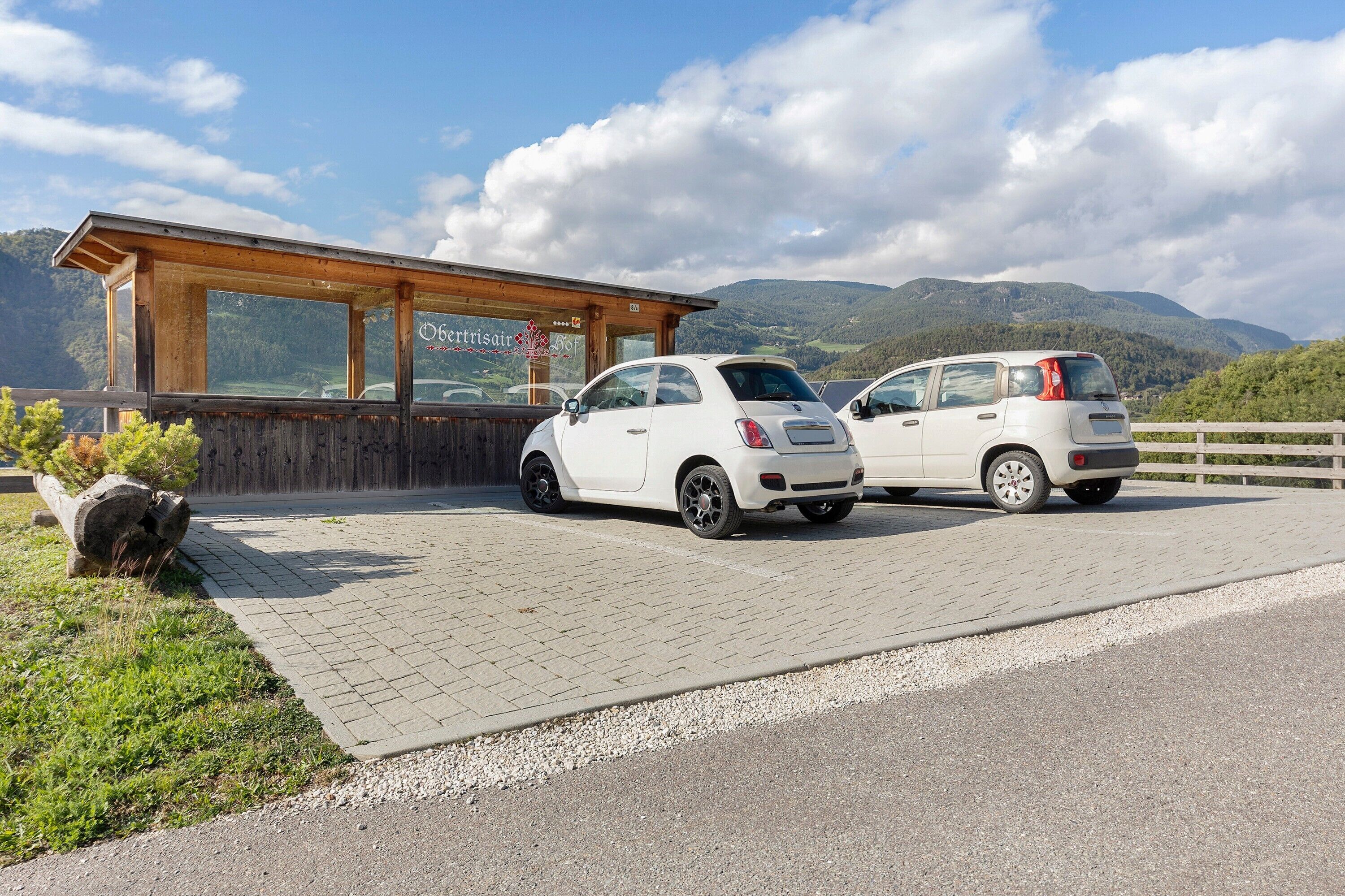 Ein Gebäude mit Holzfassade und großen Fenstern, zwei weiße Autos davor geparkt, und eine Berglandschaft im Hintergrund.